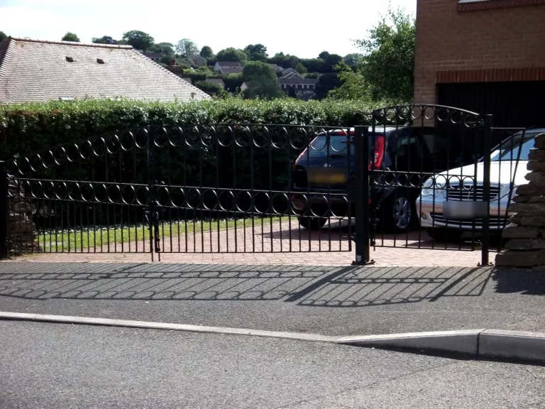 Black arched driveway gates with looped ring detail in front of a paved drive