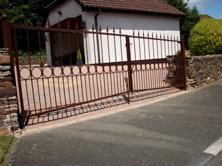 Pair of brown metal driveway gates with finials and ring band across a paved drive