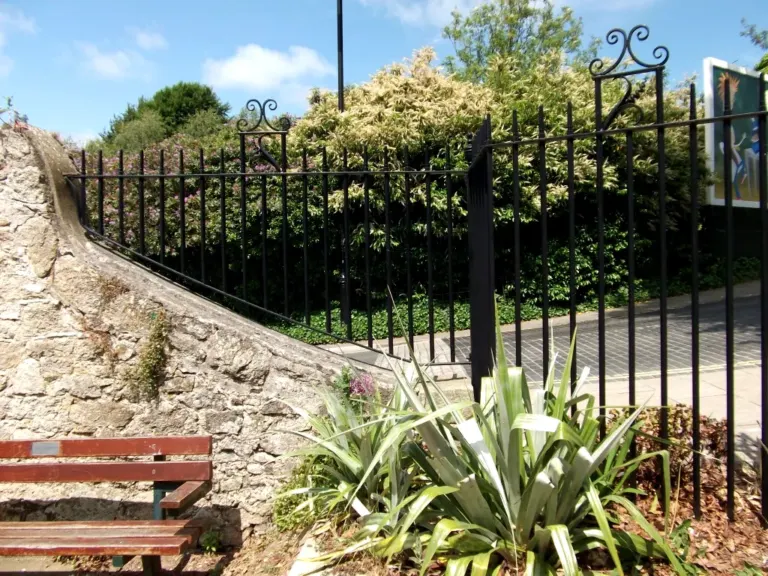 Black railings with scroll panels following a sloping stone wall beside a public garden
